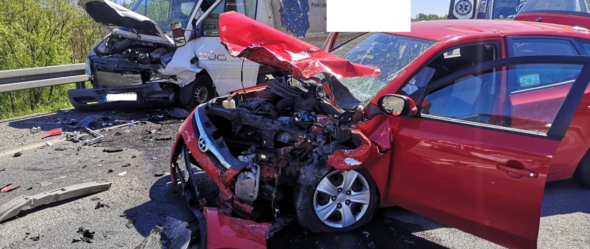 Na zdjęciu widoczny jest czerwony samochód osobowy z rozbitą komorą silnika. Maska komory silnika jest podniesiona, zderzak, błotniki przednie całkowicie zniszczone. Na drugim planie widoczny samochód dostawczy z uszkodzonym przodem - podobnie jak w samochodzie osobowym