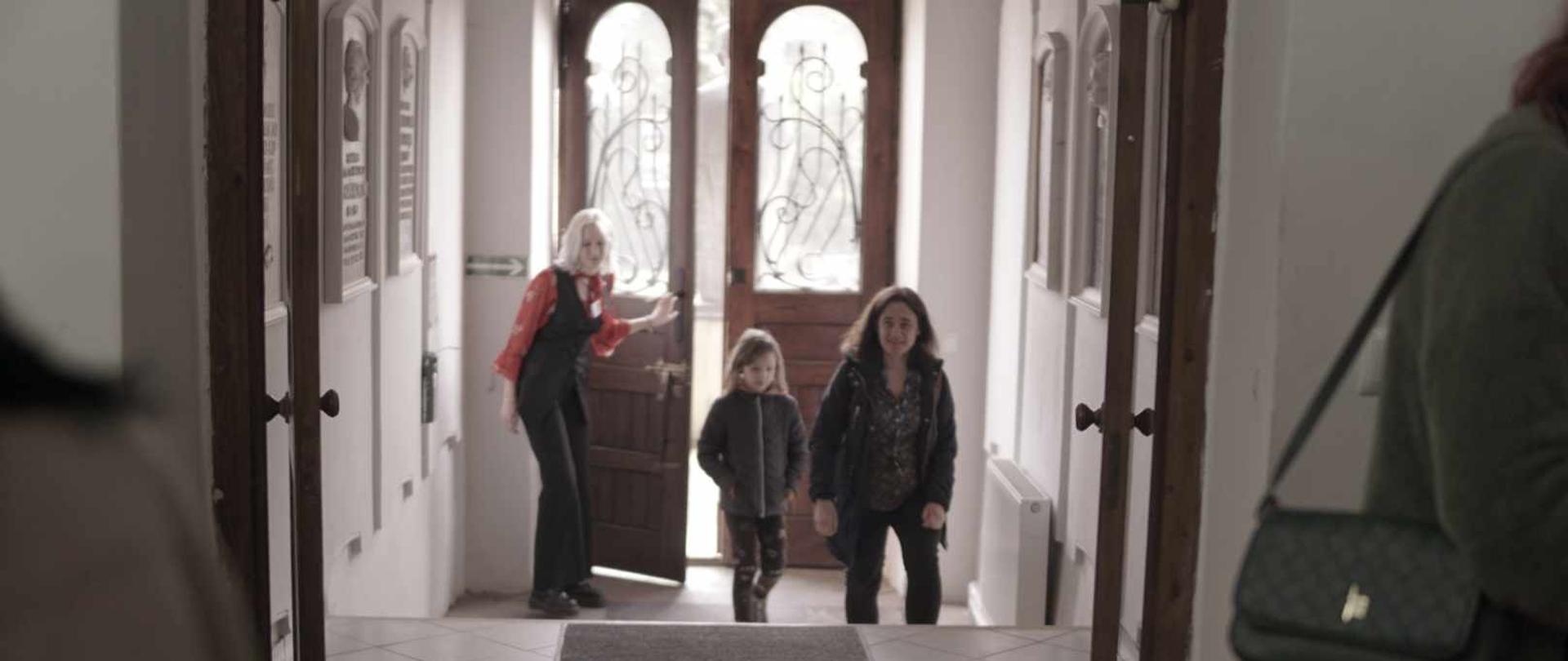 W głębi korytarza otwarte drzwi, przez które wchodzą osoby.