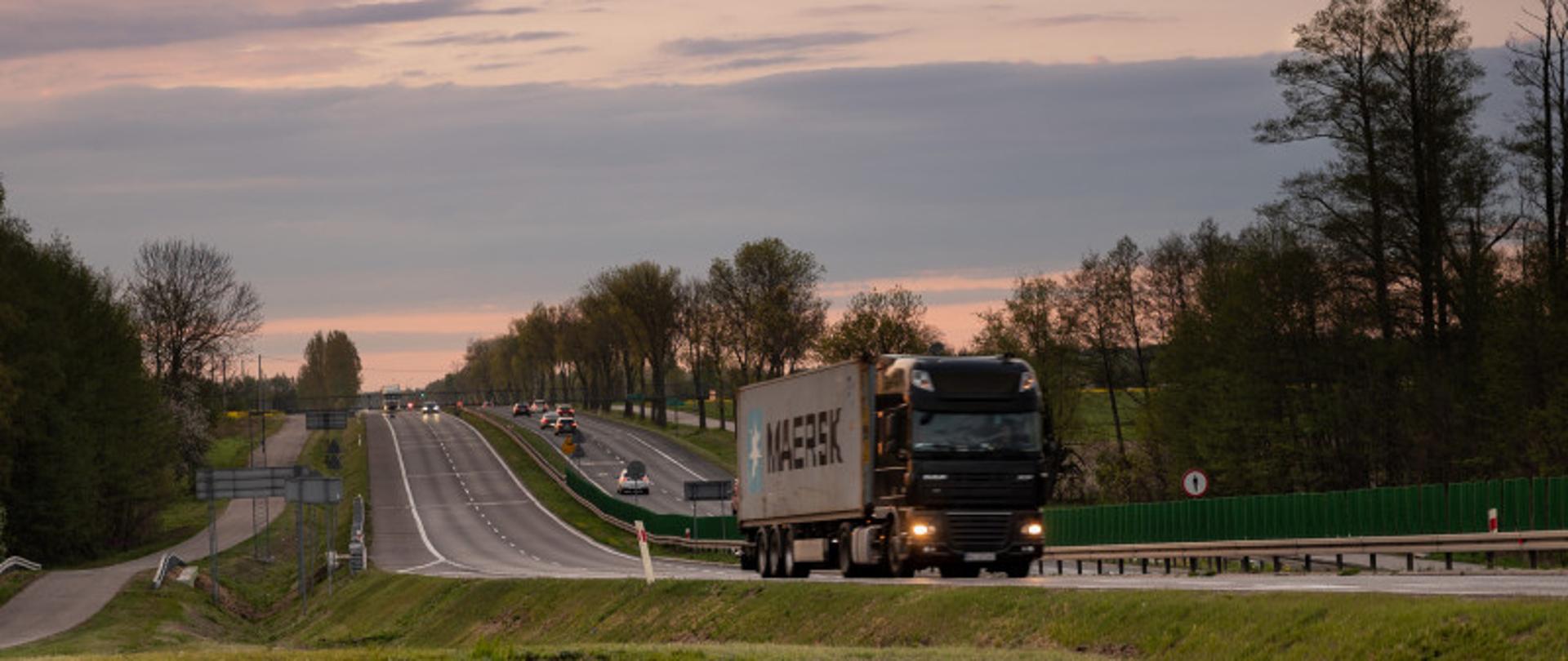 Na zdjęciu widać ciężarówkę, która porusza się po drodze dwujezdniowej. W tle widać zachód słońca.
