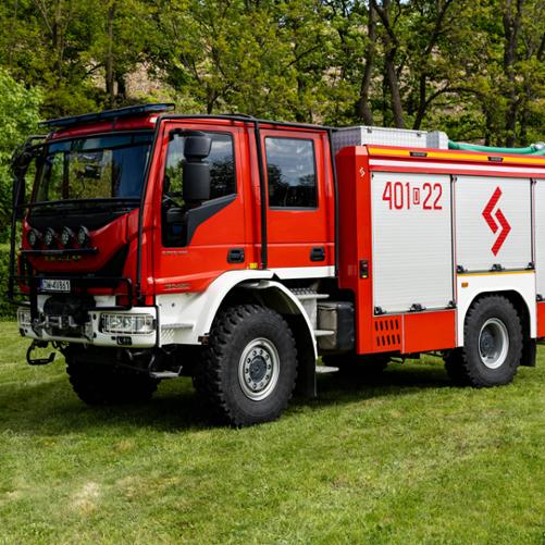 Czerwono-biały, uterenowiony samochód ratowniczo-gaśniczy marki IVECO Eurocargo na grubych, terenowych oponach. Na boku kabiny widnieje biały napis operacyjny „401[D]22”, a na żaluzji skrytki sprzętowej – czerwone, geometryczne logo. Pojazd stoi na trawiastym placu na tle zielonych drzew.