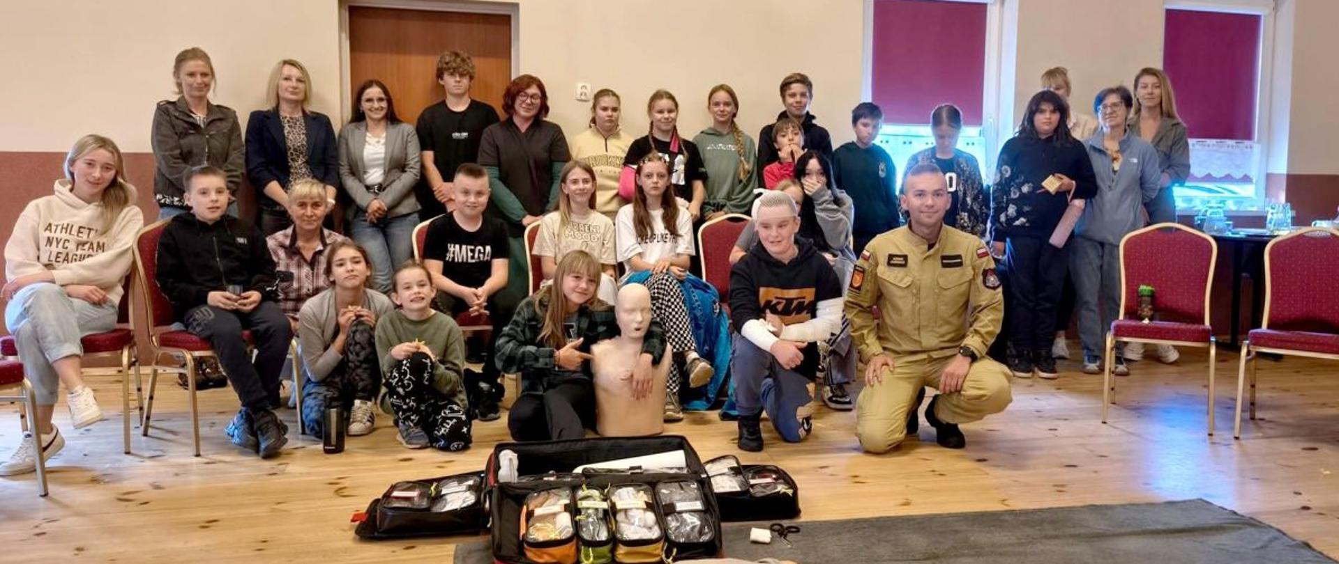 Zdjęcie przedstawia wszystkich uczestników kursu pierwszej pomocy oraz prowadzącego to szkolenie funkcjonariusza PSP. Przed grupą leży otwarta torba r1, fantom oraz szkoleniowe aed.