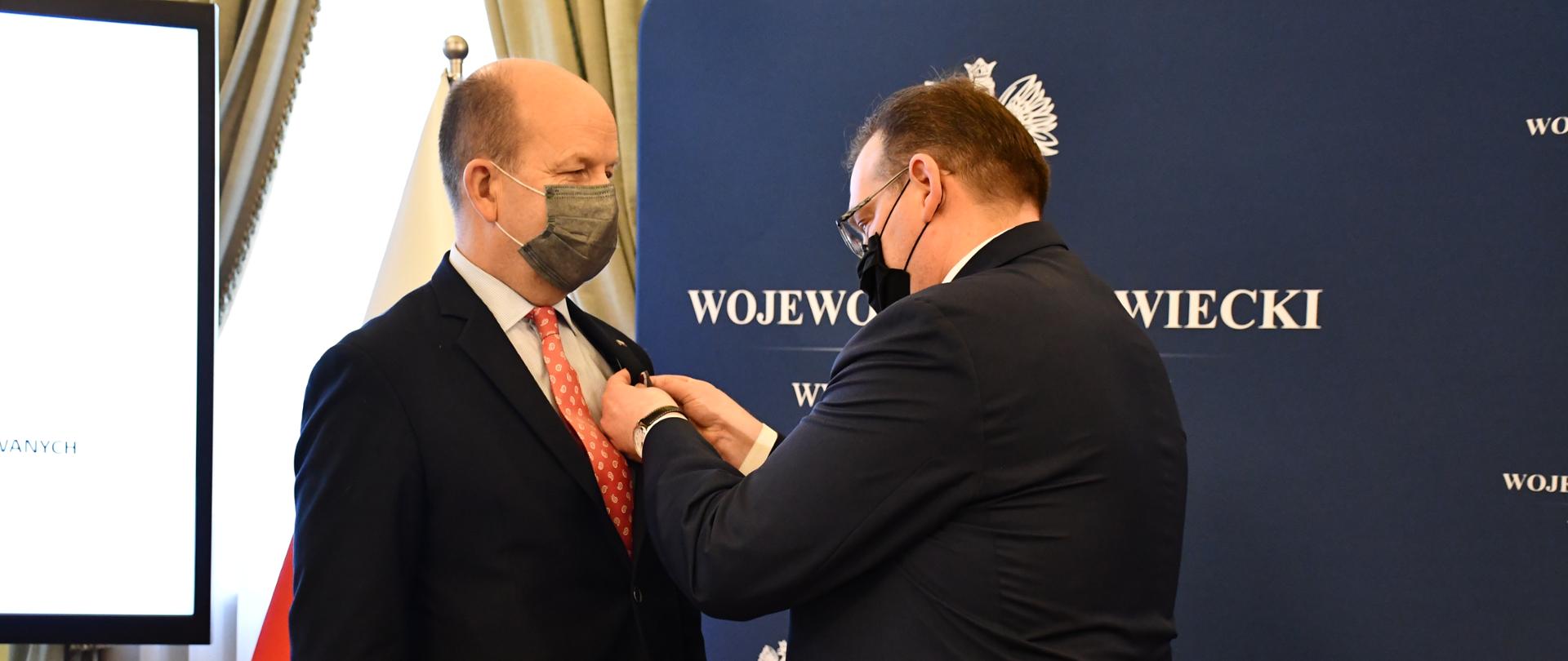 Zdjęcie przedstawia moment odznaczenia Medalem Pro Bono Poloniae. Na zdjęciu znajduje się dwóch elegancko ubranych mężczyzn. 