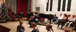 Uczeń grający na gitarze, w tle dzieci siedzące na scenie, nauczyciel i fortepian