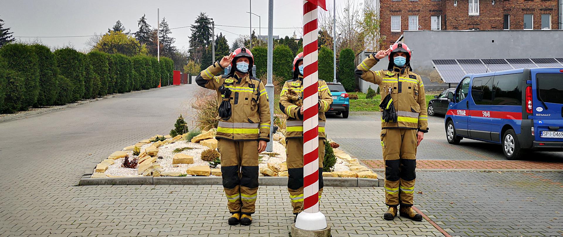Na zdjęciu widać plac przed Komendą Miejską PSP w Piekarach Śląskich, na którym 3 strażaków w ubraniach specjalnych w kolorze piaskowym i czerwonych hełmach, jako poczet flagowy, podnoszą flagę państwową na maszt z okazji Narodowego Święta Niepodległości. Z lewej strony widać rząd tuj, z prawej samochody zaparkowane na placu, a z tyłu bramę wyjazdową z Komendy i sąsiadujące z terenem komendy budynki.