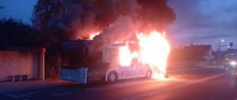 Na zdjęciu widoczny jest autobus w płomieniach. Bardziej pali się tylna część autobusu. Ogień objął większą część pojazdu. Z przodu wydobywają się kłęby czarnego dymu. Jest wcześnie rano.