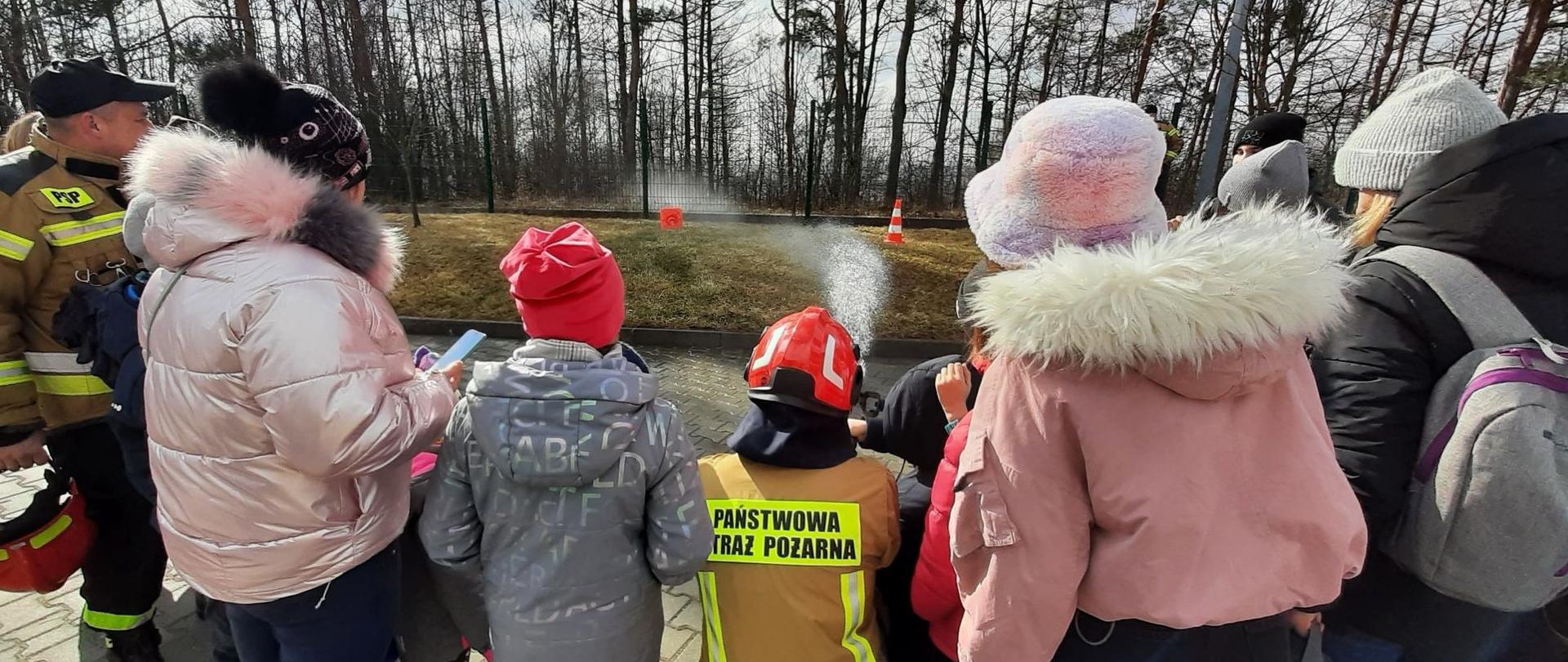 Zdjęcie przedstawia dzieci, które w asyście umundurowanego strażaka PSP podają prąd wody z szybkiego natarcia do celu jakim jest stożek sygnalizacyjny