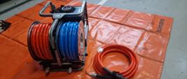 węże hydrauliczne nawinięte na zwijadło koloru czerwonego i niebieskiego obok lezy jeden krąg węza koloru czerwonego wszystko znajduje się na macie koloru pomarańczowego