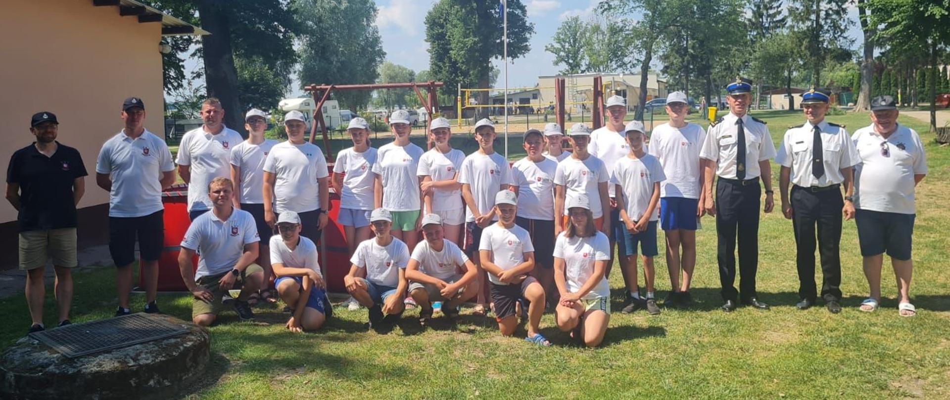 zdjęcie grupowe dzieci w białych koszulkach i kolorowych spodenkach. z boku stoją strażacy oraz mężczyźni ubrani także na sportowo