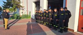Zdjęcie przedstawia strażaków stojących w dwuszeregu przed budynkiem strażnicy. Bramy garażu podniesione do góry. Z tyłu widać samochody strażackie