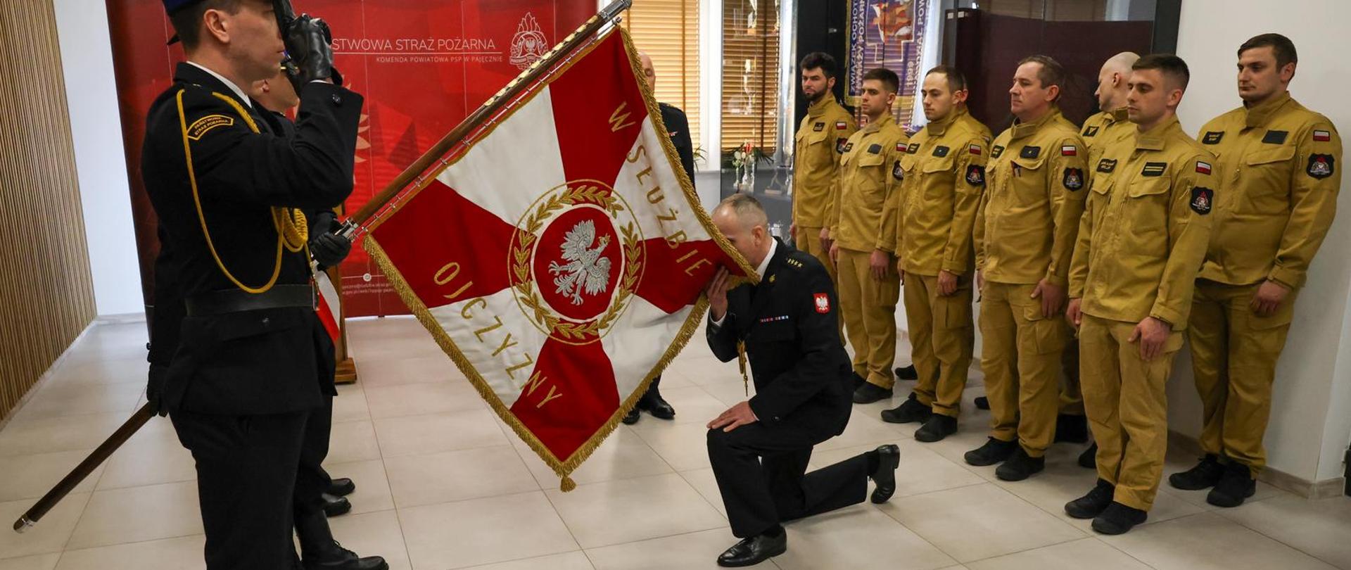 Kluczowy moment uroczystości. Oficer w ciemnym mundurze klęka na jedno kolano i całuje rąbek sztandaru, na którym widnieje napis „W SŁUŻBIE OJCZYZNY”. Wokół stoją strażacy w postawie zasadniczej, oddając honory.