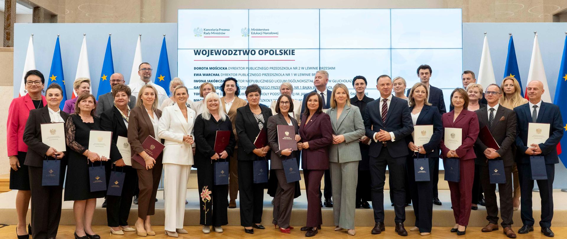 Duża grupa mężczyzn i kobiet, ubranych w eleganckie stroje, stoi na scenie, pozując do zdjęcia. Większość z nich trzyma w rękach dyplomy lub teczki. W tle widoczne są flagi Polski i Unii Europejskiej oraz ekran z napisem „WOJEWÓDZTWO OPOLSKIE”.