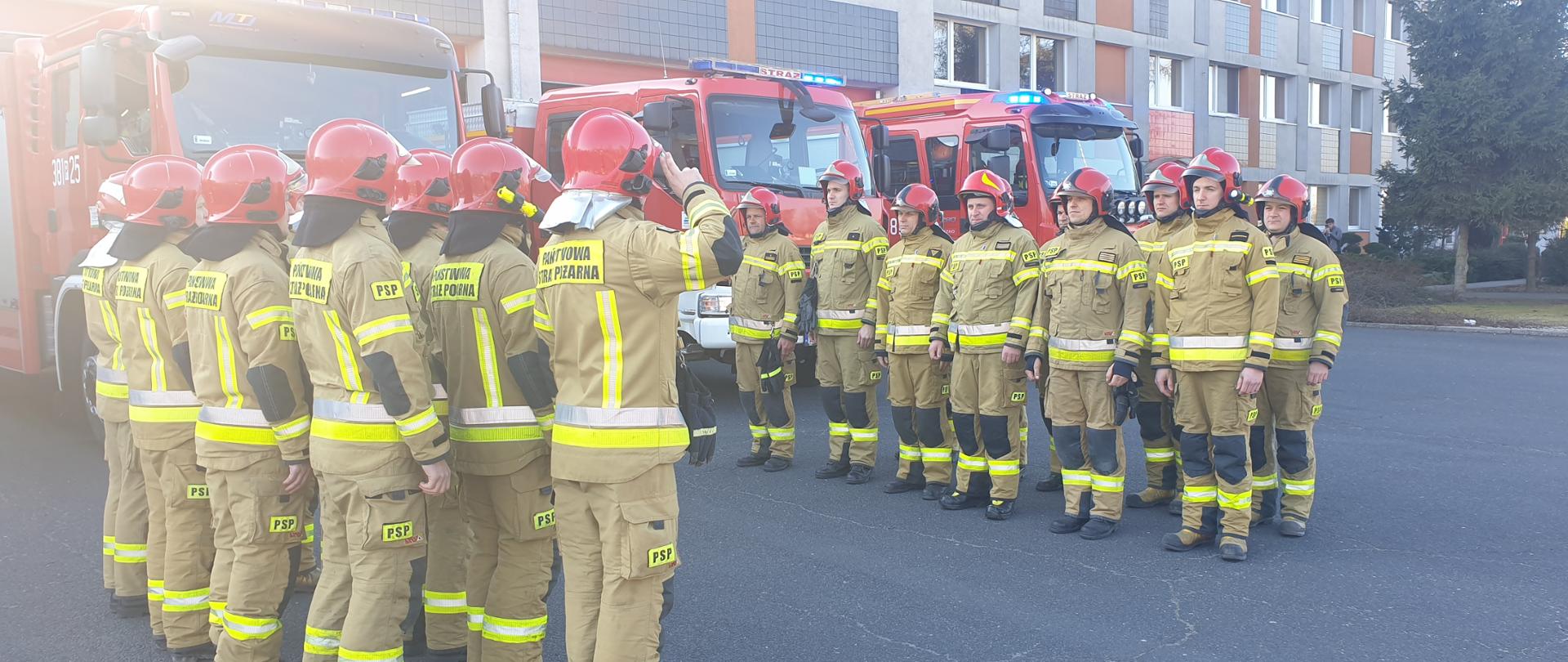 Strażacy stoją w dwóch szeregach na placu. W tle widoczna komenda i pojazdy gaśnicze.