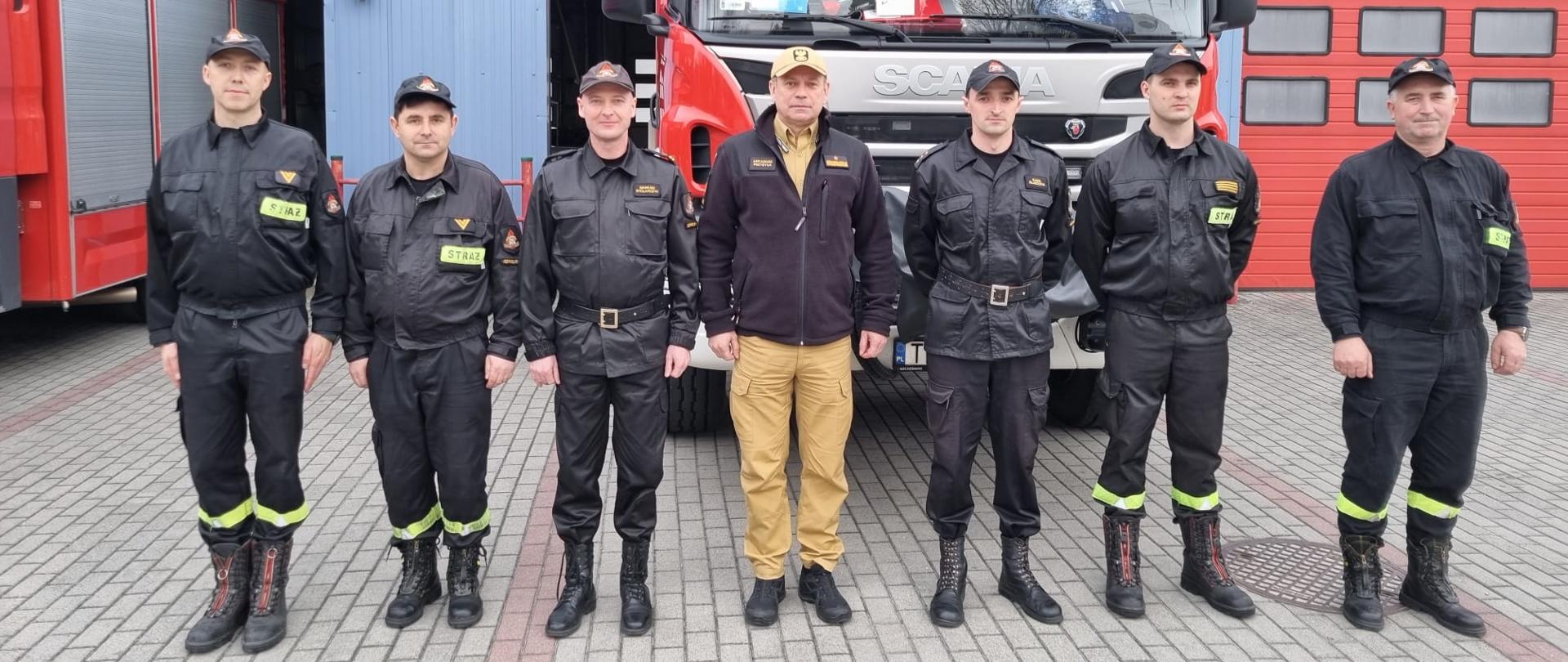Zdjęcie przedstawia zastępcę komendanta głównego Państwowej Straży Pożarnej nadbrygadiera Arkadiusza Przybyłę, komendanta powiatowego PSP w Jędrzejowie mł. bryg. Mariusza Stolarczyka oraz zmianę służbową jednostki ratowniczo-gaśniczej. Wszyscy znajdują się przed bramami garażowymi na tle samochodów ratowniczo-gaśniczych.