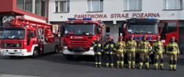 Na zdjęciu widzimy strażaków, którzy stoją na tle pojazdów pożarniczych w tle widać budynek jednostki ratowniczo gaśniczej. 