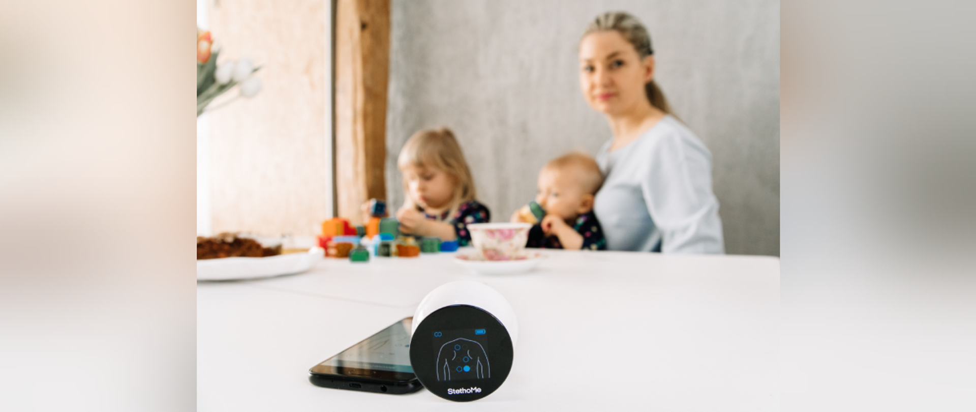 StethoMe w użyciu - urządzenie stoi na stole obok telefonu. W tle Kobieta z dwójką dzieci