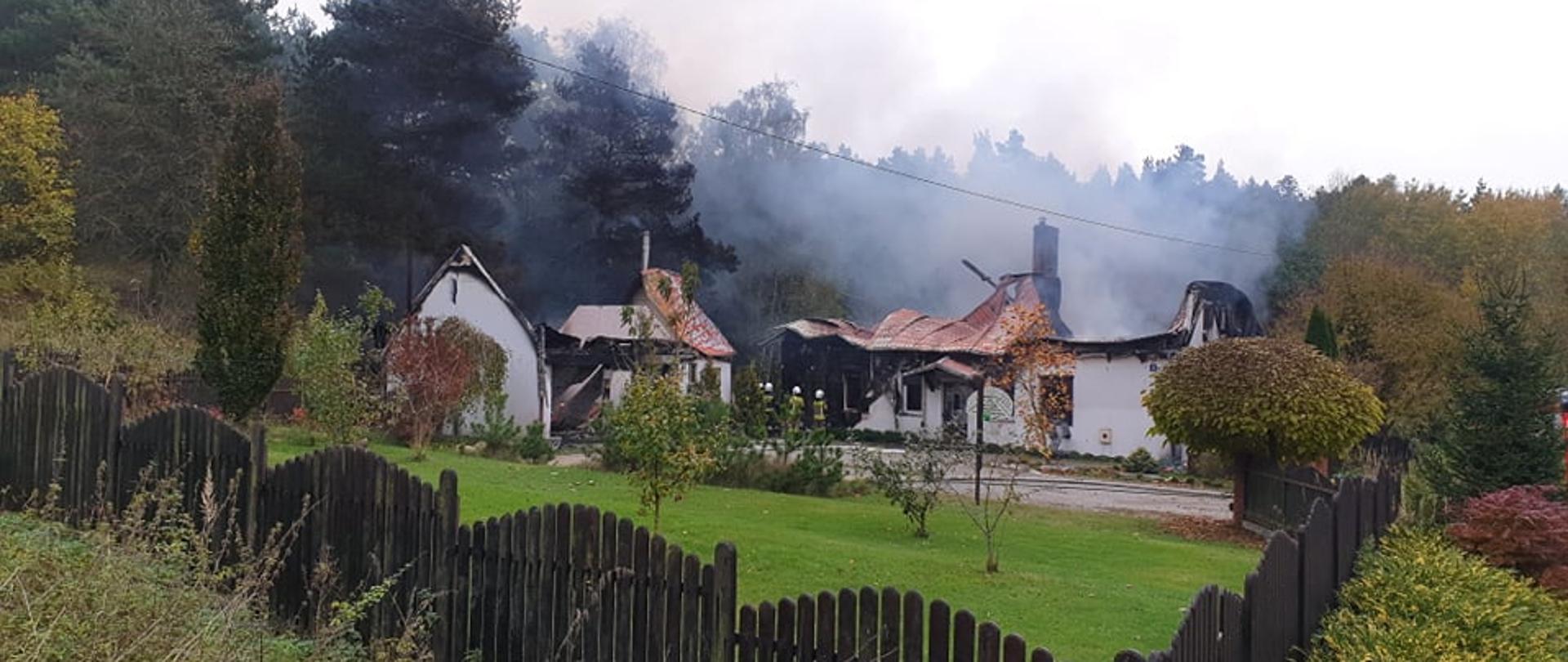 Zdjęcie przedstawia zniszczone dwa budynki po pożarze, po prawej stronie budynek mieszkalny jednorodzinny, po lewej stronie budynek garażowo-gospodarczy. Zapadnięte dachy, okopcone ściany, unoszący się dym. Posiadłość ogrodzona drewnianym płotem. W tle las.