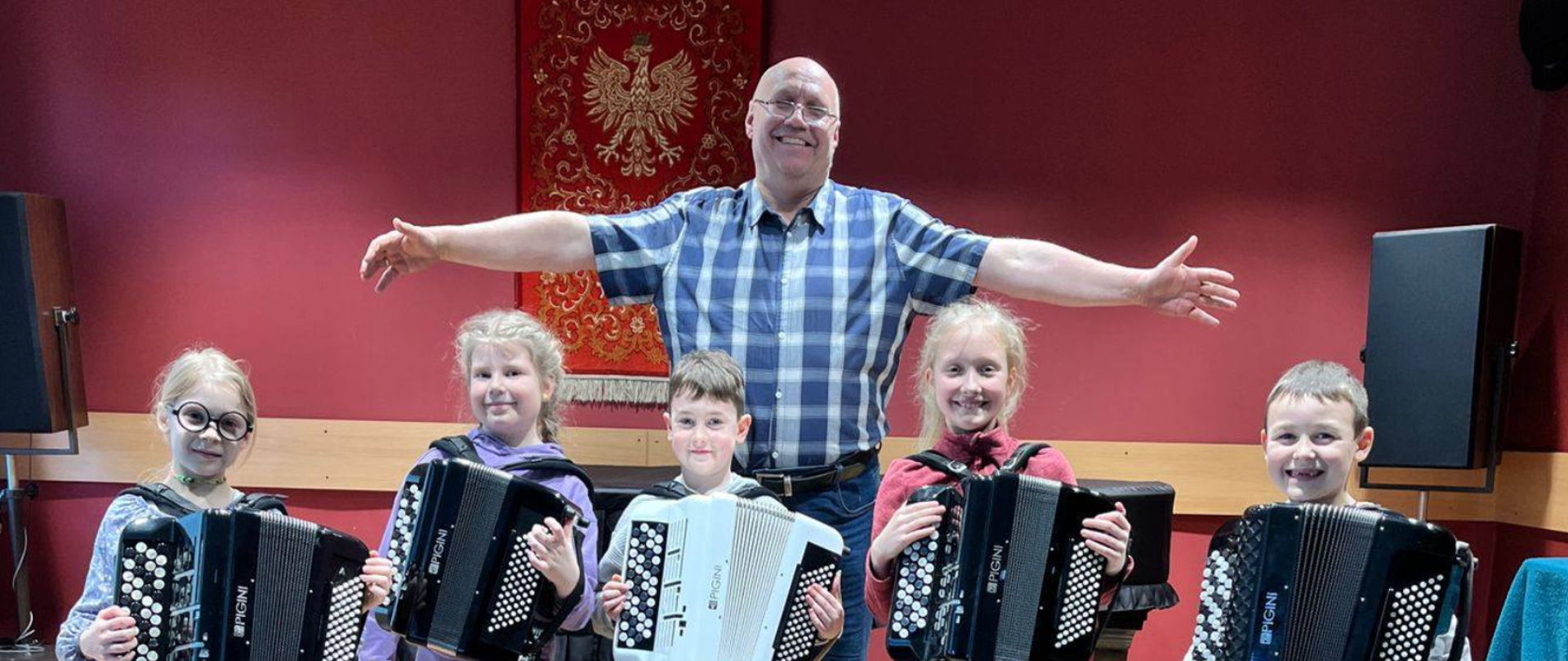 Plakat w odcieniach różu, bieli i czerni promujący koncert akordeonowy. W górnej części znajduje się logo szkoły z profilem Fryderyka Chopina. Na środku duży napis „Wiosenny Akordeon”. Po prawej stronie grafika czarnego akordeonu. Na dole plakatu znajduje się fotografia: pięcioro dzieci stojących w rzędzie z akordeonami, za nimi uśmiechnięty nauczyciel z szeroko rozłożonymi ramionami. W tle czerwone ściany sali koncertowej i godło Polski.