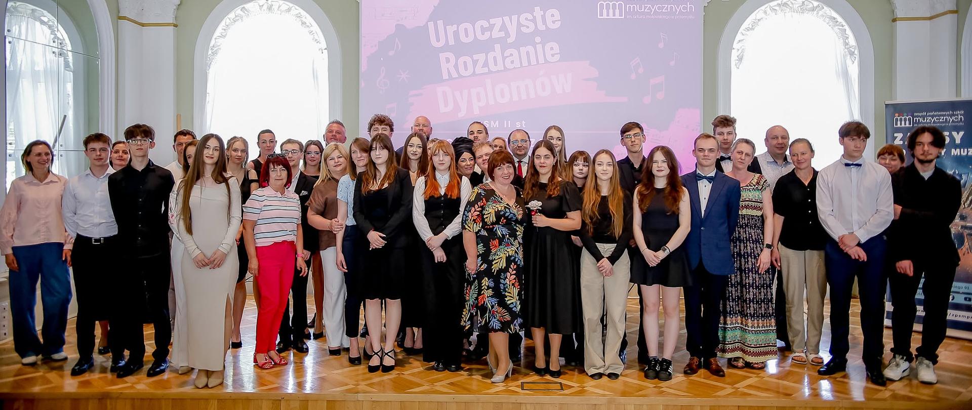 Zdjęcie przedstawia dużą grupę osób – absolwentów, nauczycieli oraz gości – pozujących do wspólnej fotografii pamiątkowej na scenie w eleganckiej, klasycystycznej sali koncertowej.