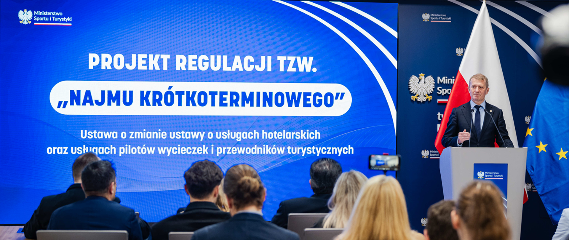 Konferencja prasowa Ireneusza Rasia ws. najmu krótkoterminowego. Minister stoi w garniturze za mównicą na polskiej flagi. Po lewej stronie ekran z tytułem konferencji