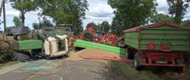 Widok na miejsce zdarzenia. Po lewej leżący traktor. Po prawej naczepa, która się nie przewróciła. Na środku wysypane zboże na drogę. Za zbożem leżąca do góry nogami naczepa.