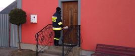 Strażacy roznoszą ulotki do gospodarstw domowych na terenie powiatu ostrowieckiego. Na zdjęciu widać strażaka przed domem mieszkalnym, który zostawia broszurę informacyjną.