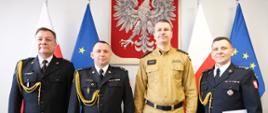 Wspólne zdjęcie oficerów Państwowej Straży Pożarnej. Stoją od lewej st. bryg. Paweł Kielar, bryg. Grzegorz Brzozowski, bryg. Grzegorz Szyszko i st. bryg. Piotr Dudek. W tle Godło Rzeczypospolitej Polskiej oraz Flagi Rzeczypospolitej Polskiej i Unii Europejskiej.