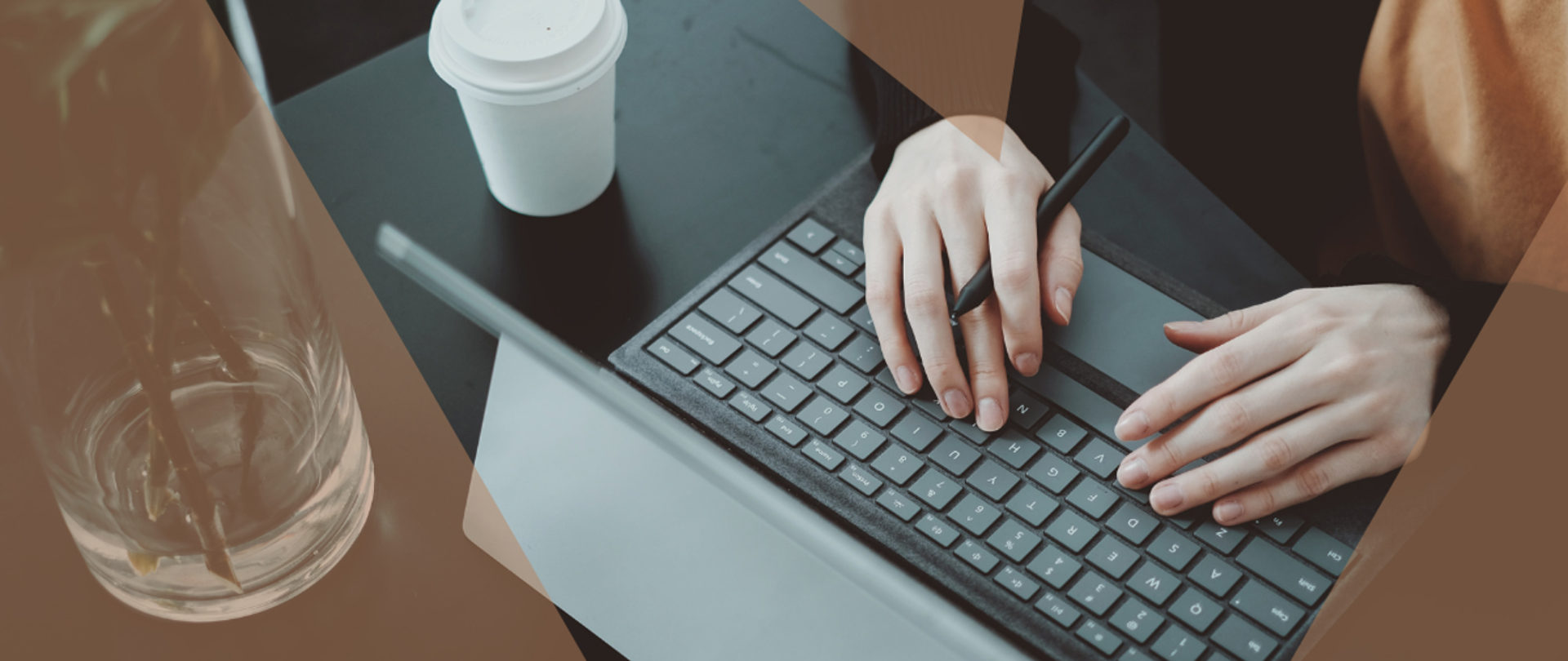 Osoba pracująca przy użyciu laptopa z długopisem w ręku. Na stole obok komputera kubek z napojem.