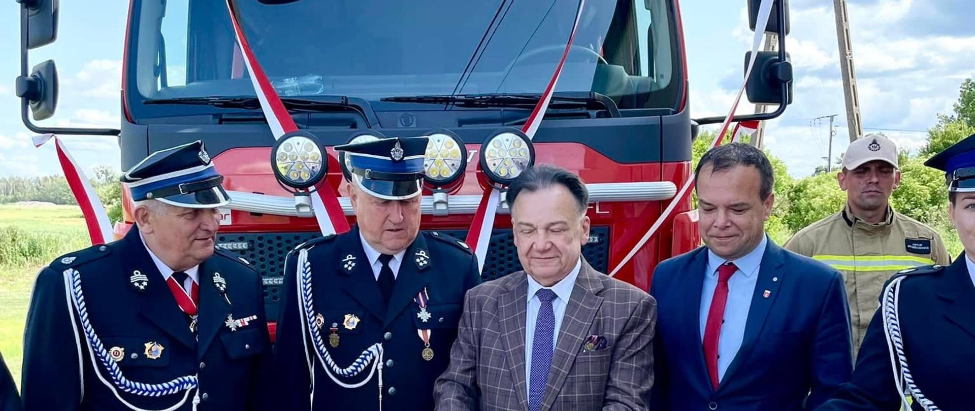 
Zdjęcie przedstawia scenę uroczystego przecięcia wstęgi z okazji przekazania nowego pojazdu strażackiego. Centralnym punktem jest duży, czerwony wóz strażacki, na którego przodzie widnieje napis "KSRG OSP BIEŻUŃ", wskazujący na jego przynależność do Krajowego Systemu Ratowniczo-Gaśniczego Ochotniczej Straży Pożarnej w Bieżuniu. Wóz jest udekorowany biało-czerwonymi wstążkami, typowymi dla takich ceremonii.