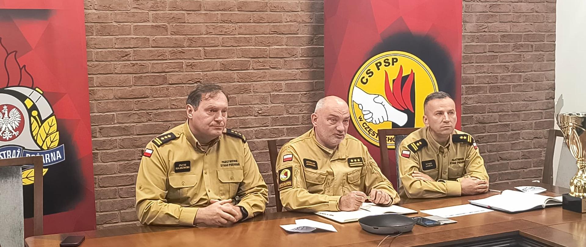 Trzech oficerów PSP siedzących za stołem konferencyjnym podczas wideokonferencji, w tle po lewej stojący baner PSP po prawej baner CS PSP