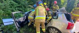 Rozbity samochód widziany z lewej strony. Dokoła widoczne sylwetki strażaków podczas próby ewakuowania osoby poszkodowanej z wraku pojazdu. W tle pojazd pożarniczy.