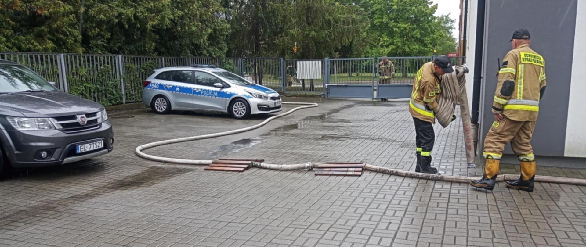 Na zdjęciu widzimy dwóch strażaków z OSP w pełnym umundurowaniu, pracujących przy rozkładaniu lub zwijaniu węża strażackiego. Działania odbywają się na brukowanym placu przy budynku. W tle stoi radiowóz policji oraz inny pojazd osobowy. Pogoda jest deszczowa — powierzchnia placu jest mokra. W tle znajduje się ogrodzenie z metalowych prętów oraz drzewa.