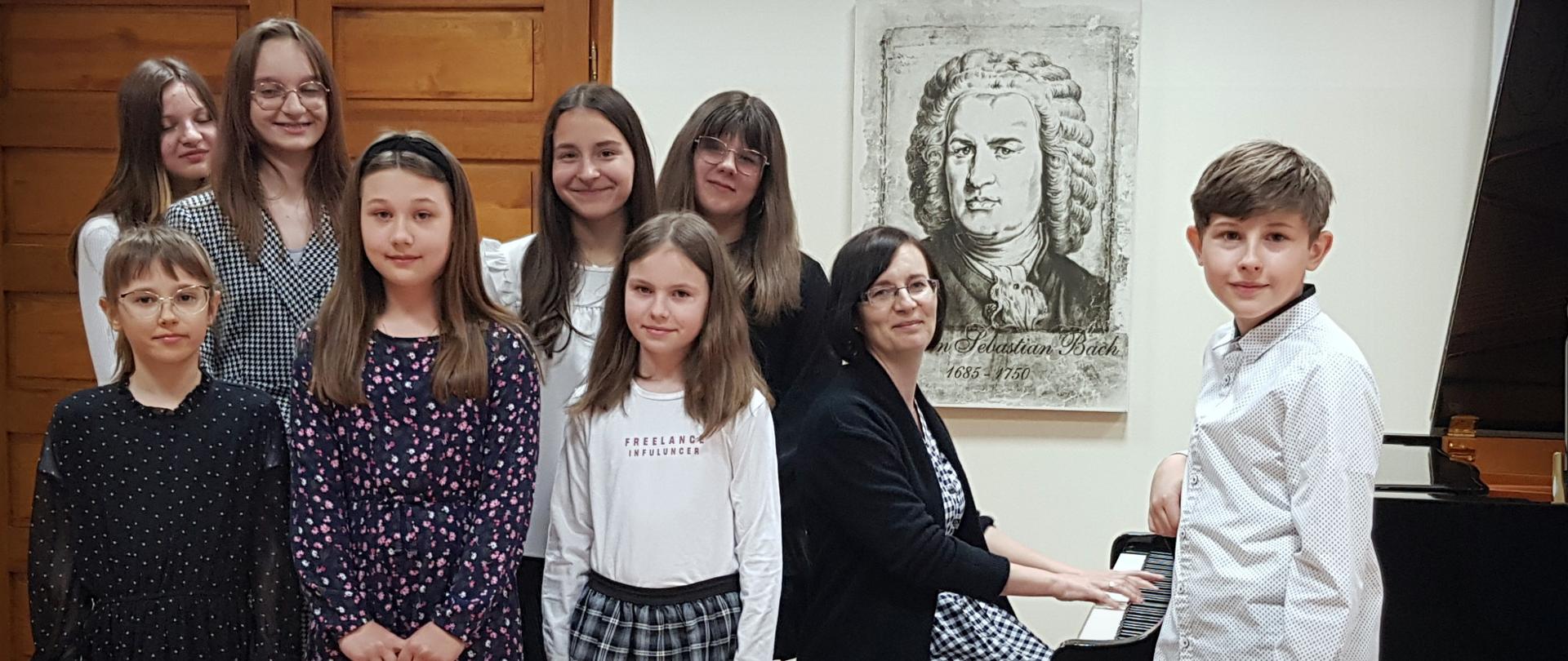 Na zdjęciu widać uczniów Pani Ewy Nowackiej stojących na scenie Auli PSM I st. w Grajewie. Przy fortepianie siedzi nauczycielka Pani Ewa Nowacka. Uczniowie i ich nauczycielka ubrani są w kolory biało, czarno, granatowe. W tle drewniane drzwi, oraz grafika z J. S. Bachem.