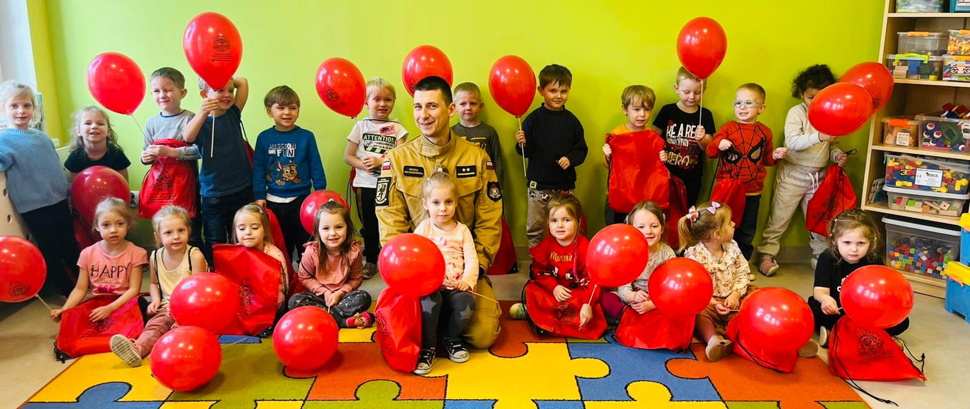 Na zdjęciu widać rozbawioną grupę przedszkolaków z balonami, po środku strażak prowadzący zajęcia.