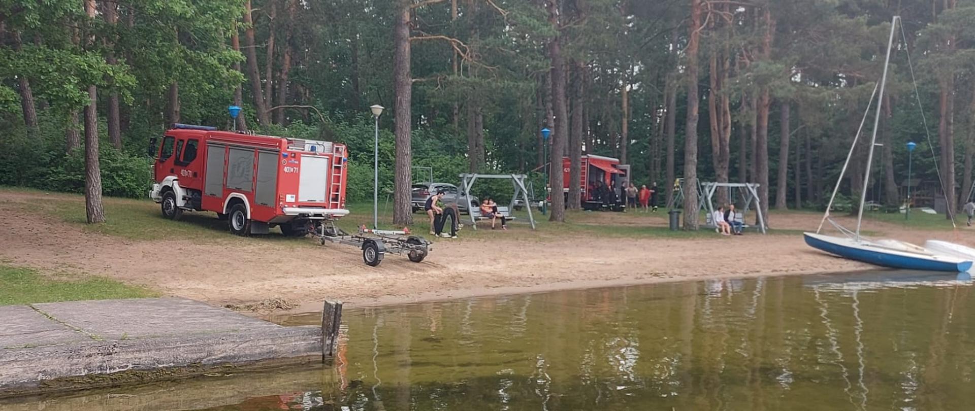 Na pierwszym planie woda i kawałek pomostu, W tle, na plaży, stoją pojazdy straży pożarnej.