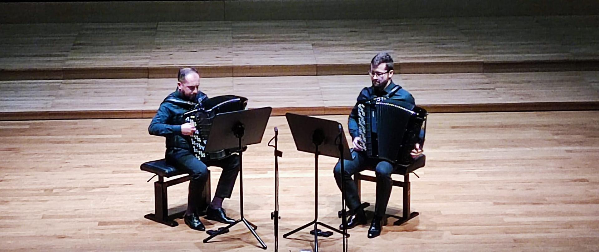 Dwóch mężczyzn zwróconych lekko do siebie siedzi na krzesłach na estradzie sali koncertowej i grają na akordeonach. Przed nimi stoją pulpity.