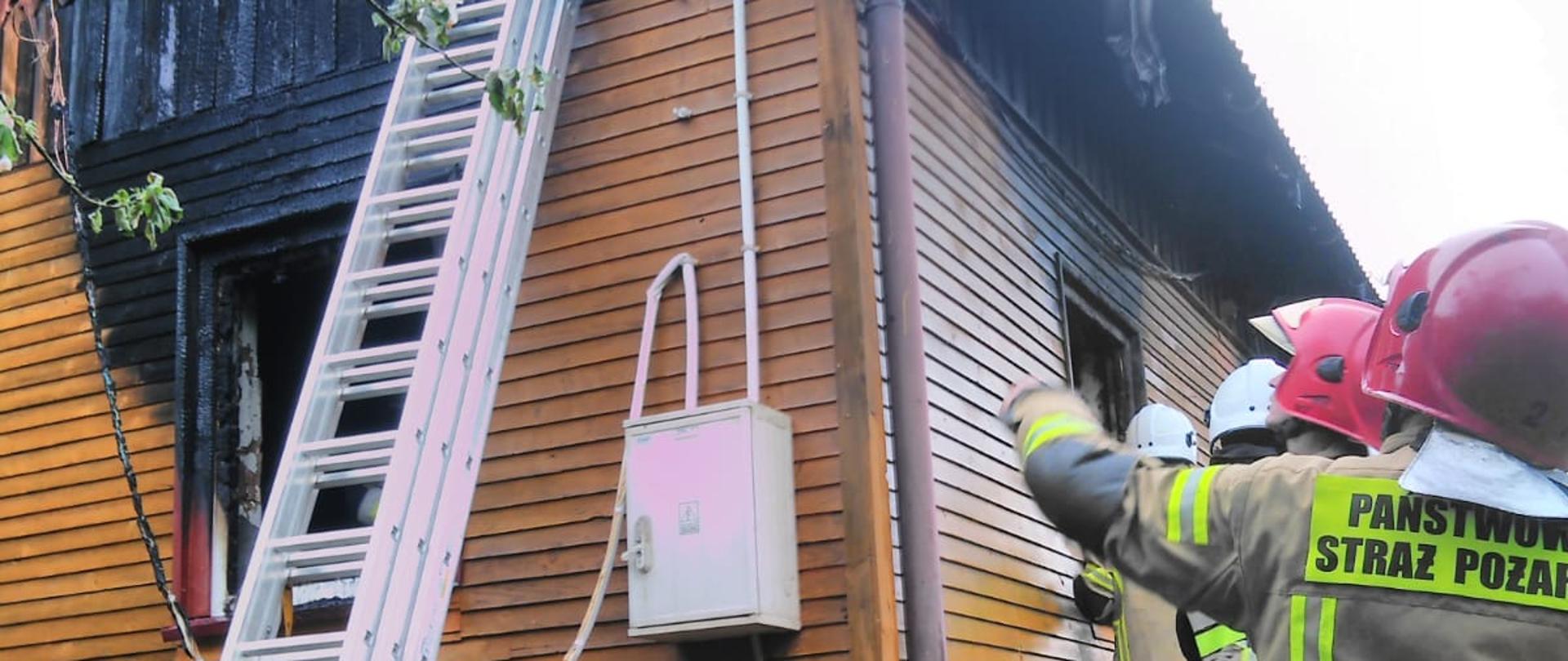 Kolorowa fotografia przedstawia na głównym planie widok narożnika budynku mieszkalnego. Budynek na podmurówce koloru ceglanego, ściany obłożone brązową deską szalunkową. Widoczna część narożna dachu, z elementami deskowania. Do budynku przystawiona trzy przęsłowa drabina aluminiowa. Widoczne opalenia obicia ściany nad wypalonym, pustym oknie. Na ścianie budynku, przy jego narożniku biała skrzynka z przyłączami elektrycznymi. Po prawej stronie częściowo widoczne postacie kilku strażaków w jasnych ubraniach bojowych i czerwonych hełmach, jeden wskazuje ręką i patrzy na budynek. Pod oknem przepalone kawałki desek. U góry w lewym narożniku fotografii widoczna zielona gałąź drzewa.