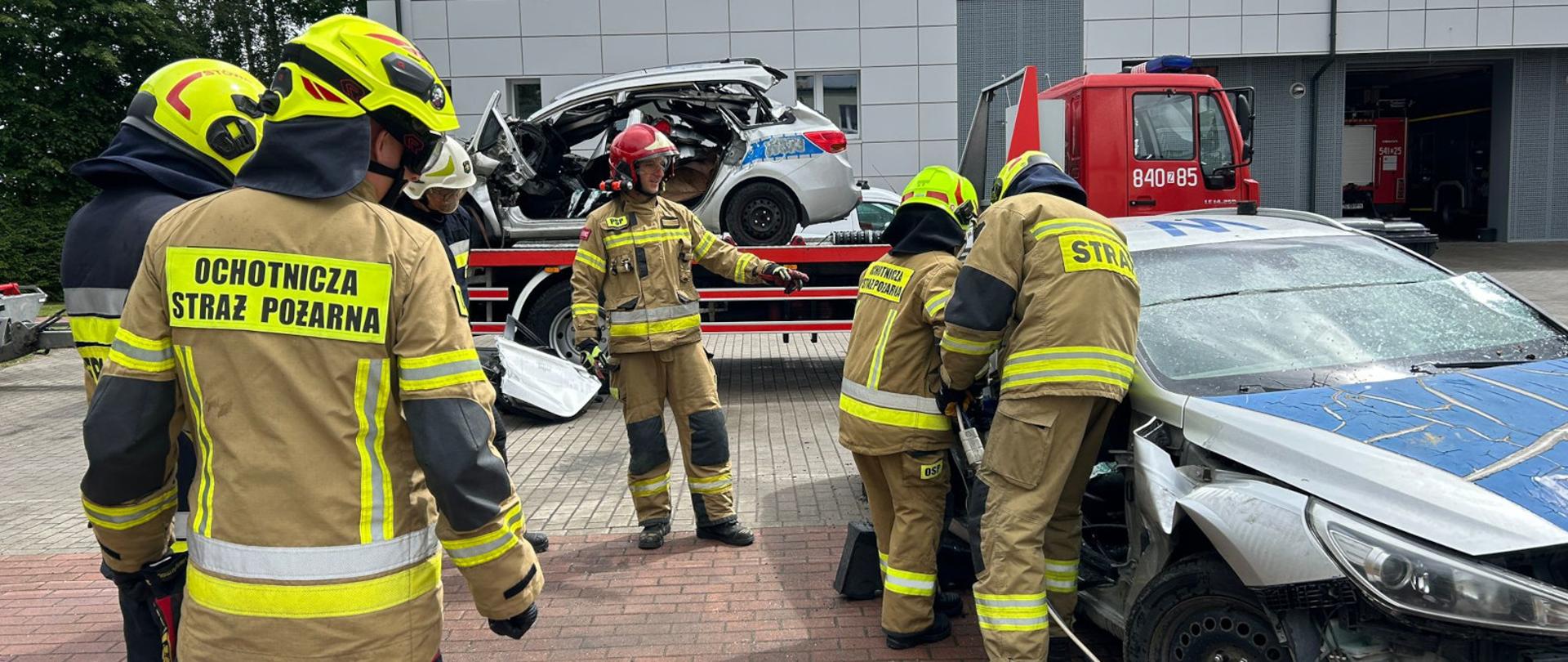 Uczestnicy egzaminu kończącego szkolenie podstawowe strażaków ratowników OSP realizują zadanie z ratownictwa technicznego przy uszkodzonym samochodzie osobowym. Dwóch strażaków pracuje przy pojeździe z wykorzystaniem sprzętu hydraulicznego, podczas gdy pozostali obserwują przebieg ćwiczenia i oczekują na swoją kolej. Nad prawidłowym wykonaniem zadania czuwa egzaminator. W tle widoczne są wrak pojazdu ustawiony na lawecie oraz samochód pożarniczy. Zdjęcie dokumentuje praktyczną część egzaminu sprawdzającą umiejętności działań ratowniczych podczas wypadków drogowych.