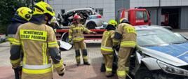 Uczestnicy egzaminu kończącego szkolenie podstawowe strażaków ratowników OSP realizują zadanie z ratownictwa technicznego przy uszkodzonym samochodzie osobowym. Dwóch strażaków pracuje przy pojeździe z wykorzystaniem sprzętu hydraulicznego, podczas gdy pozostali obserwują przebieg ćwiczenia i oczekują na swoją kolej. Nad prawidłowym wykonaniem zadania czuwa egzaminator. W tle widoczne są wrak pojazdu ustawiony na lawecie oraz samochód pożarniczy. Zdjęcie dokumentuje praktyczną część egzaminu sprawdzającą umiejętności działań ratowniczych podczas wypadków drogowych.