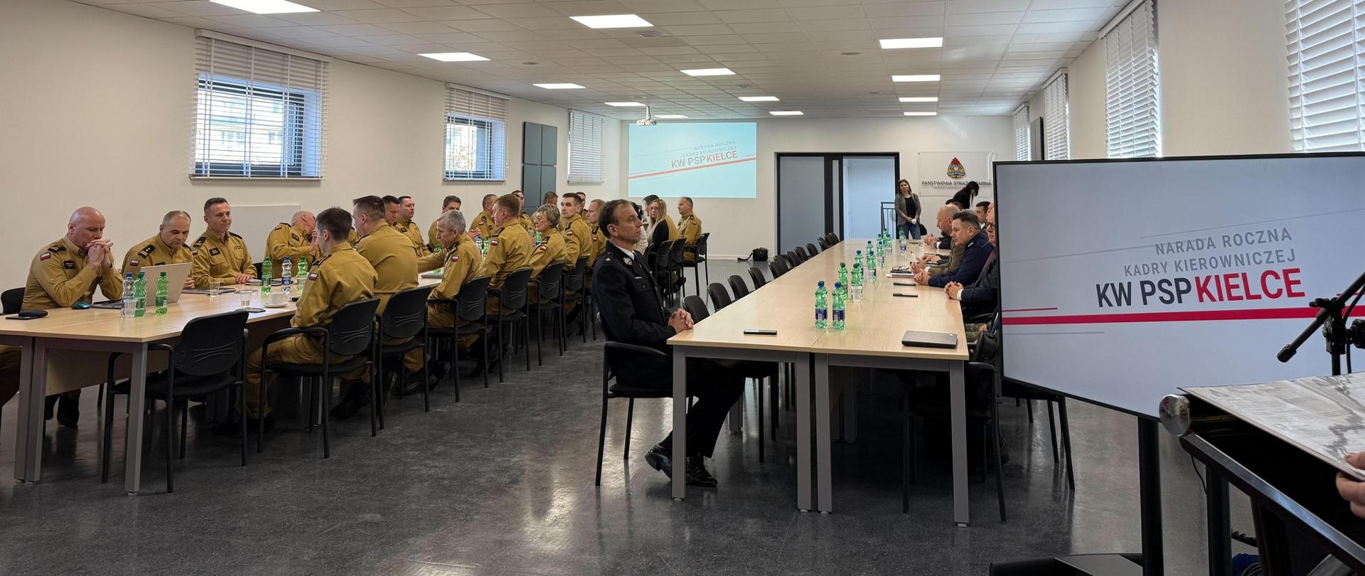 Widok sali podczas narady rocznej KW PSP w Kielcach. Na ekranie wyświetlony jest tytuł prezentacji „Narada roczna kadry kierowniczej KW PSP Kielce”. Uczestnicy siedzą przy stołach.