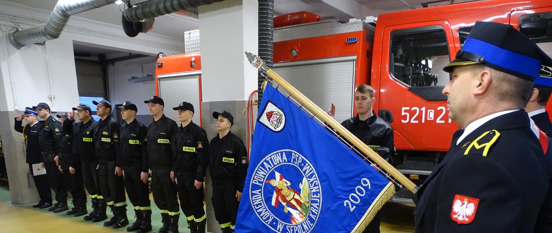 Zdjęcie wykonano w budynku Komendy Powiatowej Państwowej Straży Pożarnej w Sępólnie Krajeńskim w pomieszczeniu garaży. Zmiana służbowa stoją na baczność ubrani w mundury koszarowe. Poczet sztandarowy również uczestniczy w ceremonii, strażacy z pocztu sztandarowego są stoją ubrani w mundury wyjściowe. w tle samochód ratowniczo- gaśniczy. 