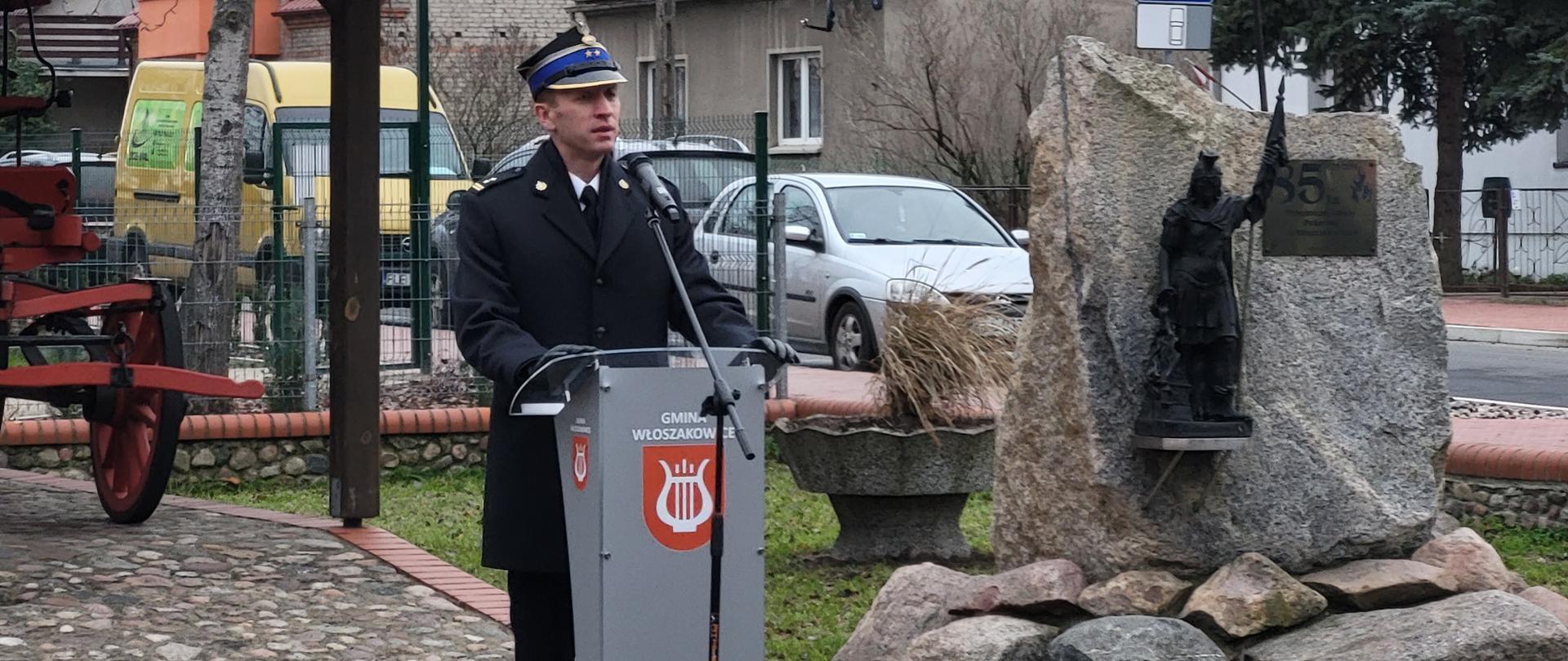 strażak w rogatywce i płaszczu stoi za mównicą, obok duzy kamieńz figurą sw. Floriana oraz stara pompa strażacka