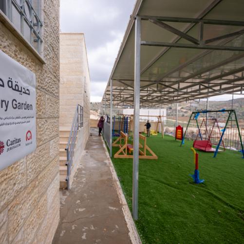 Na dalszym planie dwoje dorosłych osób stoi na zadaszonym i ogrodzonym placu zabaw dla dzieci z niepełnosprawnościami