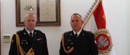 dwóch strażaków w ciemnych mundurach ze sznurem stoi obok siebie, w tle sztandar