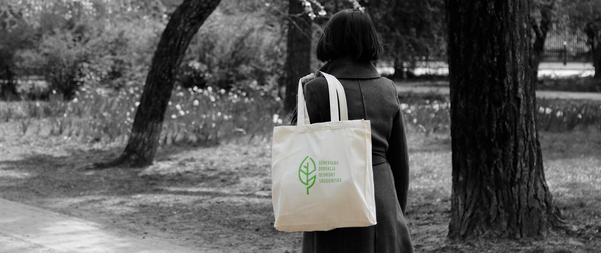 Fotografia w parku. Widać pnie drzew. Kobieta ma przerzuconą przez ramię ciemną torbę z reprodukcją obrazu oraz logotypem (biały listek) i napisem Generalna Dyrekcja Ochrony Środowiska.