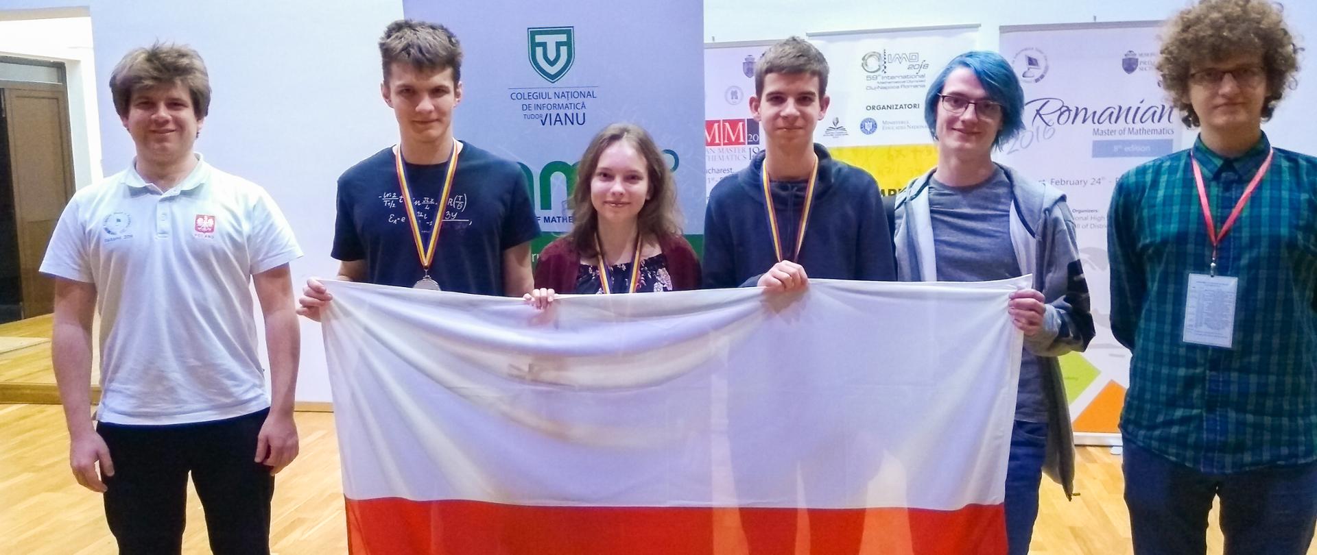 Zdjęcie przedstawia polskich uczniów uczestniczących w 12 Mistrzostwach Rumunii w Matematyce oraz ich opiekunów. Uczniowie trzymają w dłoniach biało-czerwoną flagę polskich. 