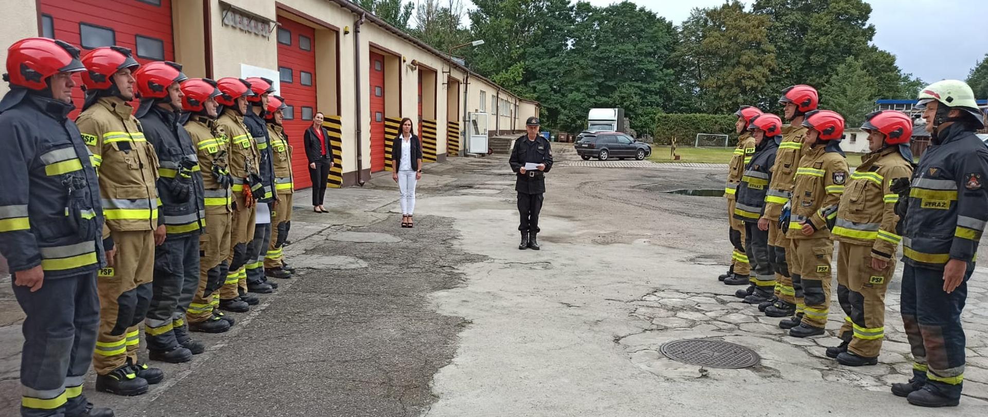 Zdjęcie przedstawia strażaków stojących w szeregu ubranych w piaskowe i czarne mundury oraz czerwone hełmy przed nimi stoją natomiast w szeregu strażacy w czarnych i piaskowych ubraniach specjalnych i hełmach, za nimi znajdują się czerwone bramy garażowe. Pomiędzy nimi znajduje się osoba trzymająca dokumenty w ręku.