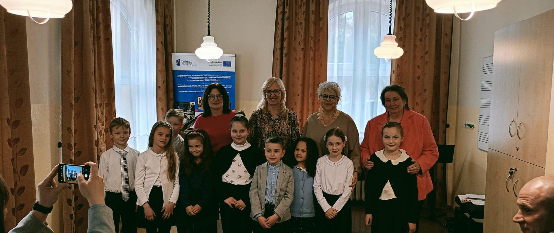 Zdjęcie grupowe. W sali stoją uczniowie grający na audycji, za nimi nauczycielki klasy fortepianu. Za nimi dwa okna przysłonięte firanami i zasłonami oraz baner unijny. Z sufitu zwisają zapalone lampy. Z prawej strony na ścianie tablica w pięciolinię oraz dwudrzwiowa szafa.