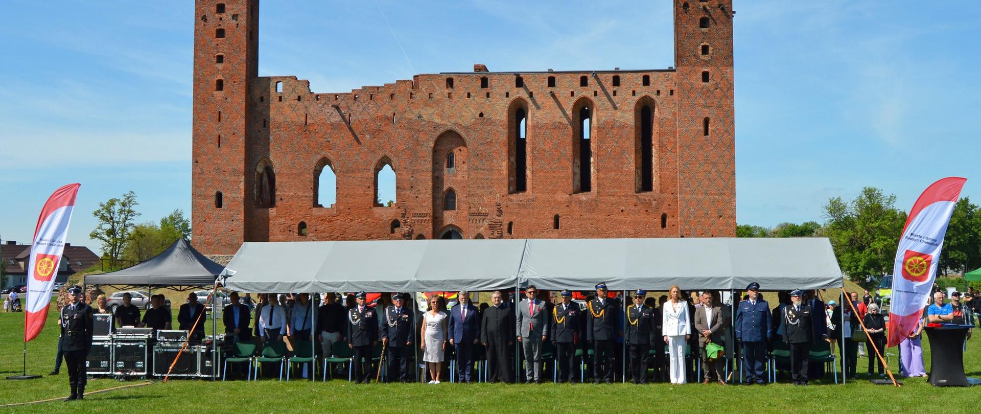Uroczysta zbiórka strażaków i osób cywilnych na terenie zielonym przed gotyckim zamkiem krzyżackim