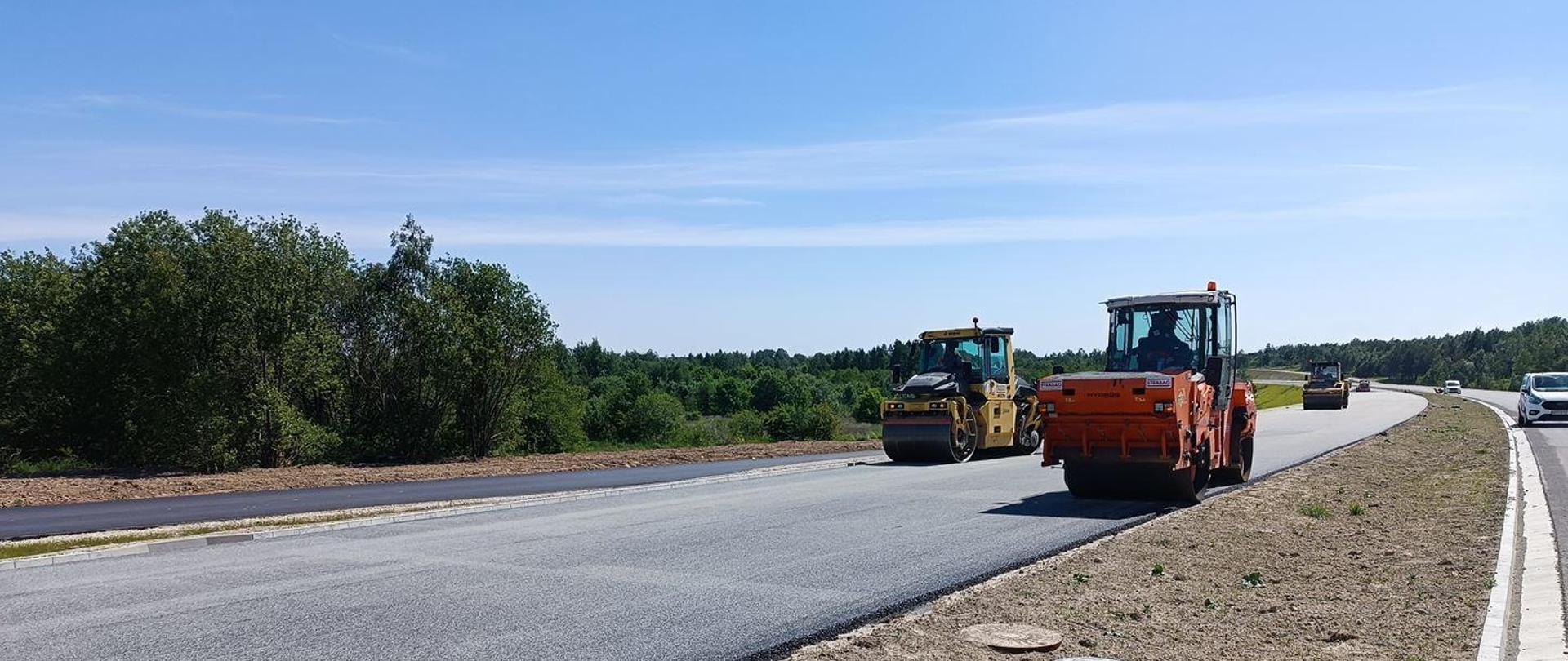 Roboty bitumiczne - układanie nawierzchni asfaltowej na jezdni budowanej dwujezdniowej drogi,. Trzy walce drogowe, w oddali samochody osobowe. Wzdłuż drogi zielone zarośla.
