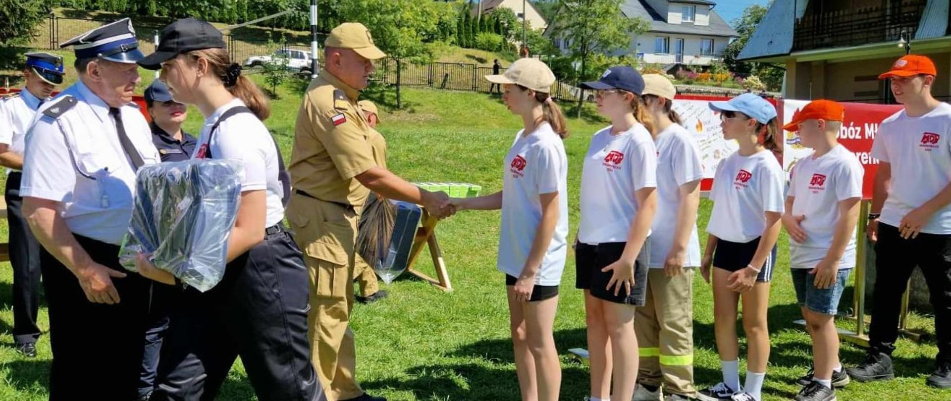 Zdjęcie zrobione na zewnątrz, na terenie ośrodka wypoczynkowego. Druh ochotnik podaje dłoń strażakowi z Młodzieżowej Drużyny Pożarniczej. Za nimi stoi oficer Państwowej Straży Pożarnej w stopniu generała. 